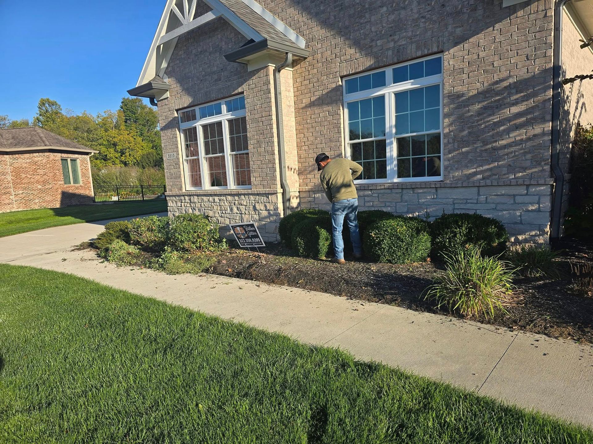 Person trimming bushes near a house with a stone facade. Sidewalk and lawn in the foreground; another brick house in the background.