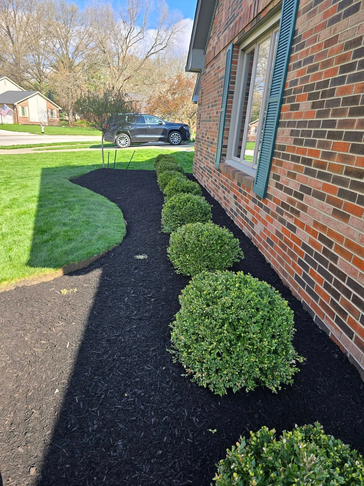 Well-manicured yard with black mulch, green shrubs, and a brick house. A car is parked on the street.