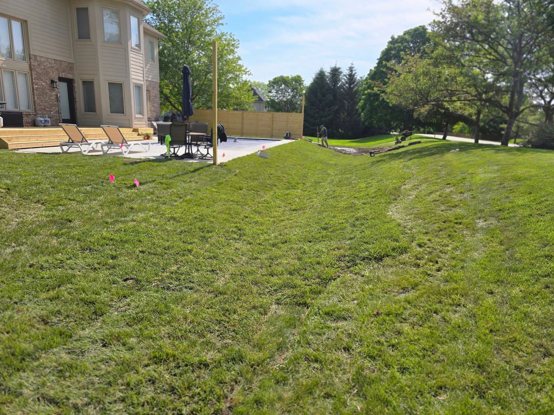 Green lawn slopes toward a patio near a beige house with a wooden deck and a brown fence.