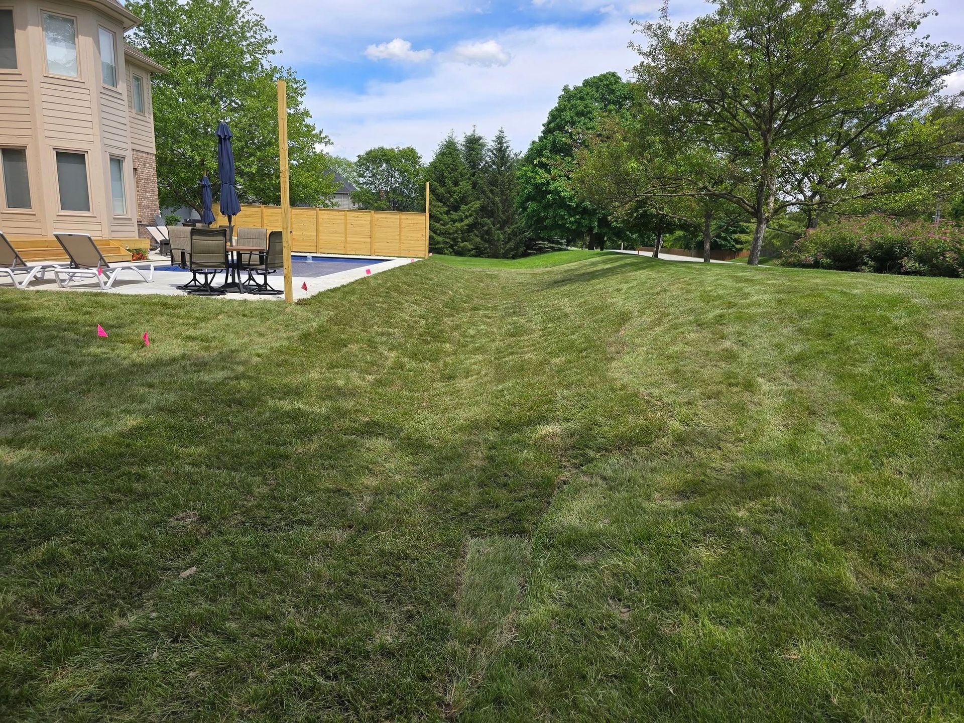 Green lawn with a house, patio, and trees under a partly cloudy sky.