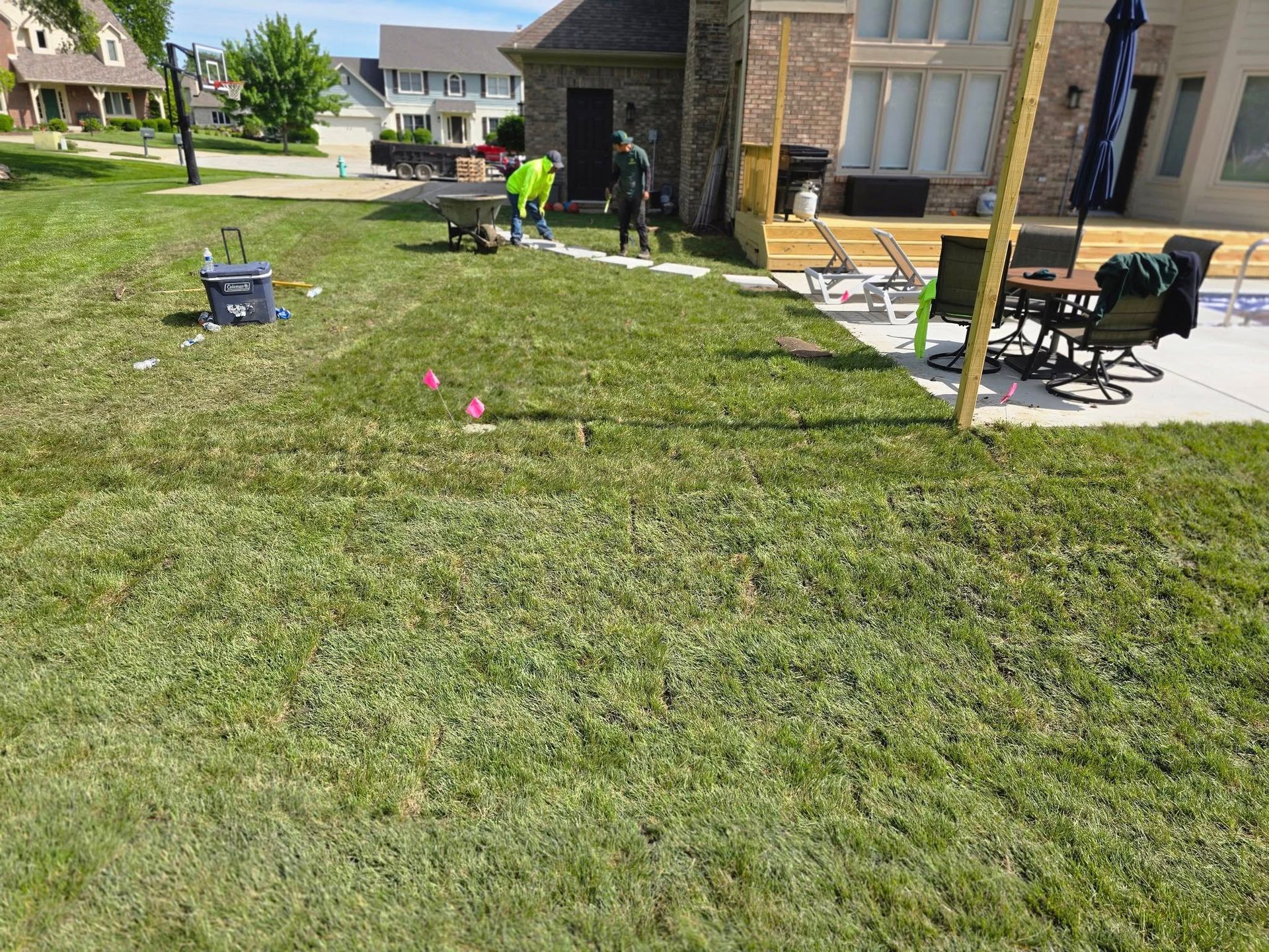 Workers installing stone edging near a house deck. Green grass, wheelbarrow, and pink flags visible.