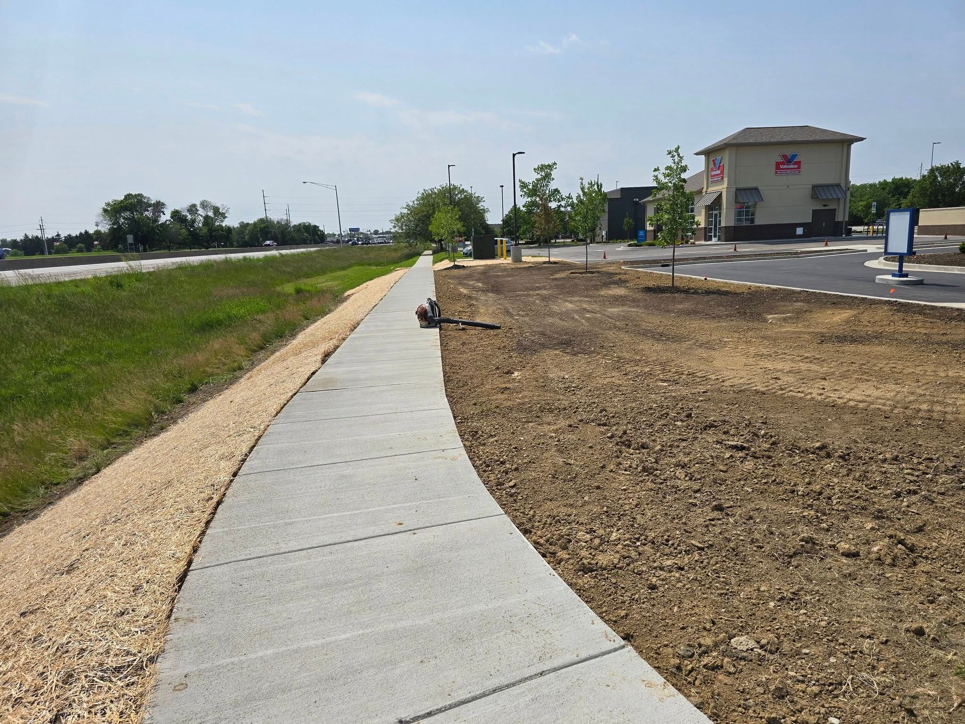 Sidewalk curves past a grassy area and dirt, leading to a building with a red logo on a sunny day.