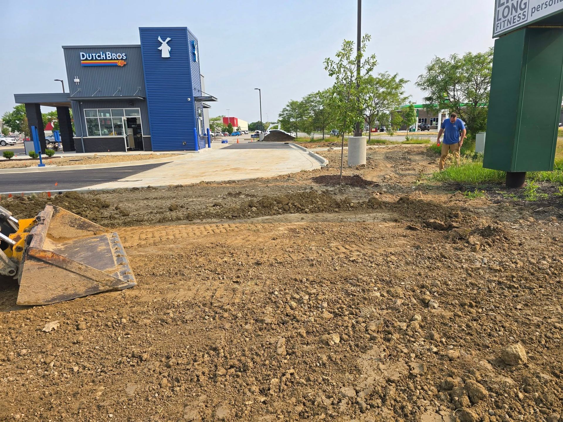 Construction site next to a blue Dutch Bros coffee shop. Earth-moving equipment and a worker visible.