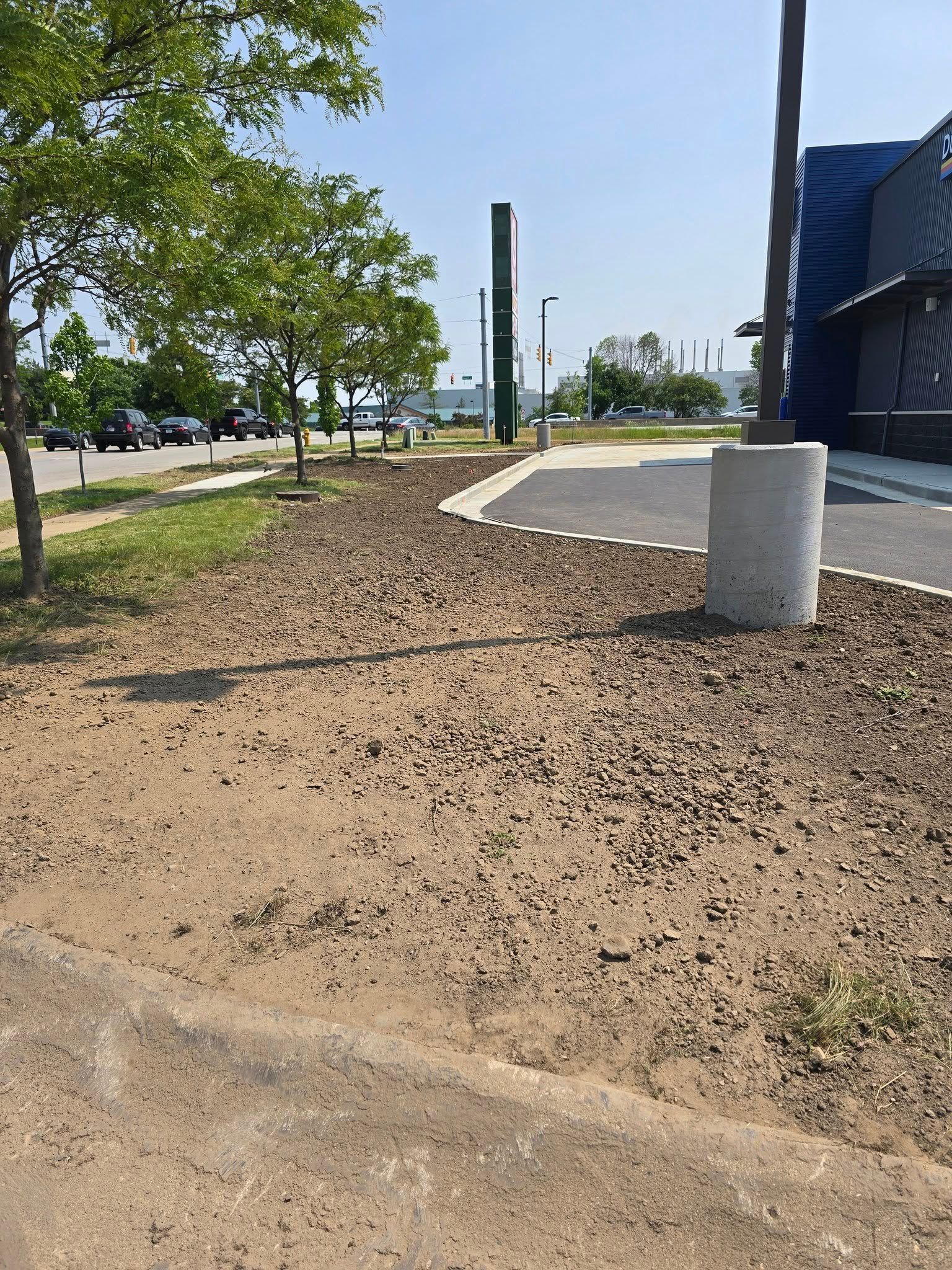 Gravel area next to a paved path with trees and a building in the background on a sunny day.
