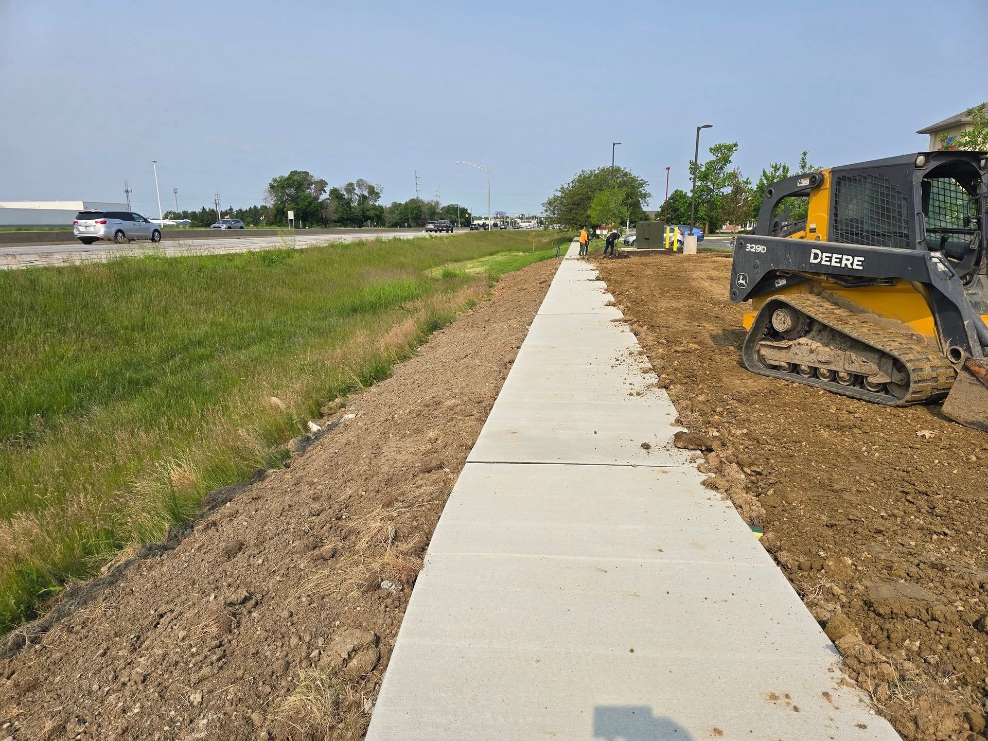 Sidewalk construction next to a grassy area with a skid steer on a sunny day.