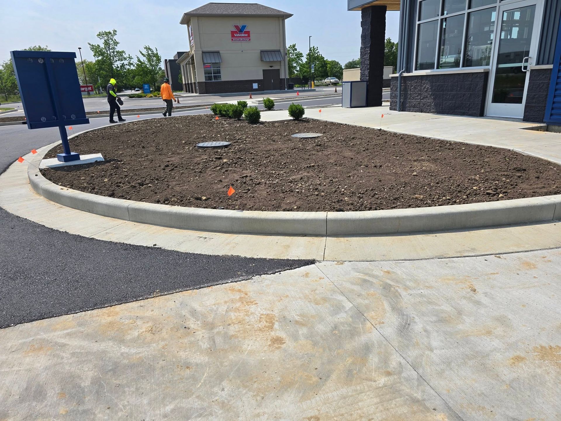 Newly planted flower bed with concrete curb, asphalt and concrete driveways, business in background.