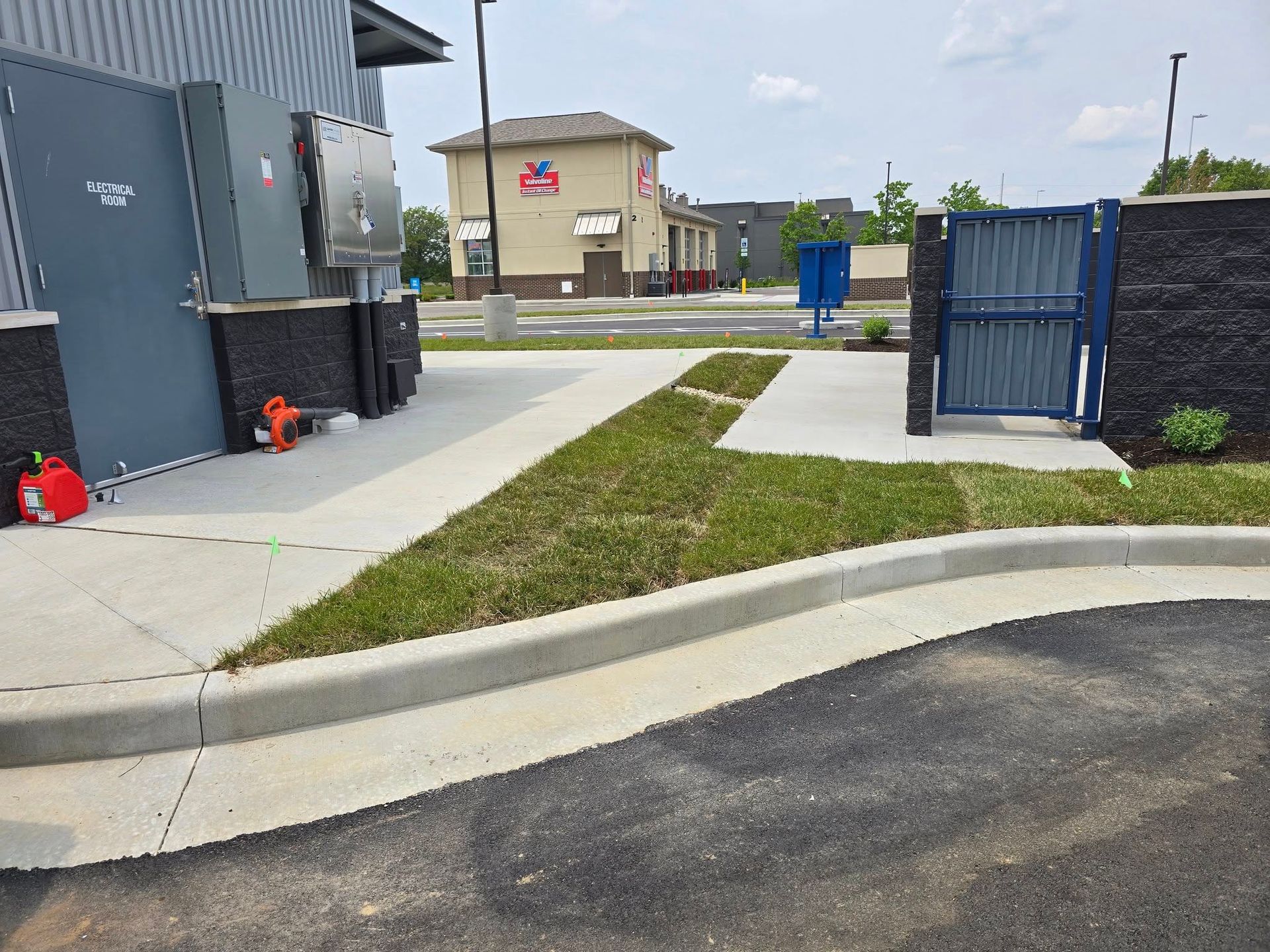 Sidewalk with curb, grass, and a gate in front of a building; a second building is visible in the background.