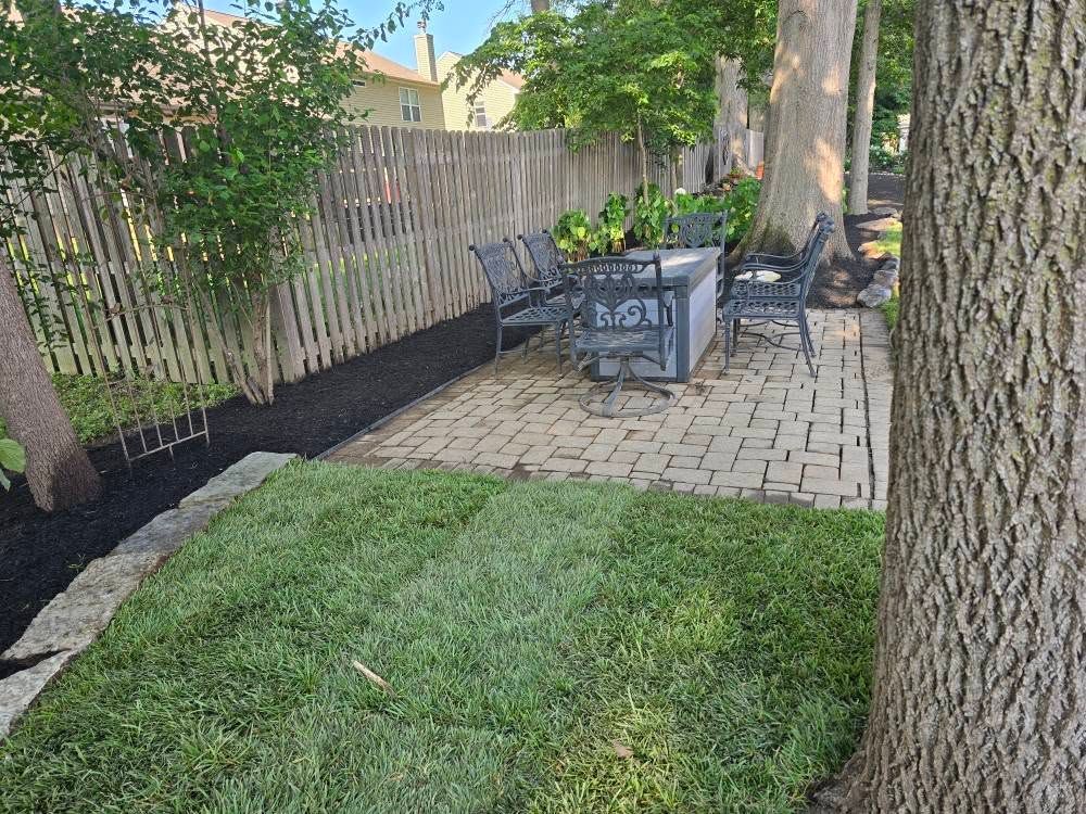 Patio with brick pavers, fire pit, and seating; lawn in foreground, wooden fence and trees.