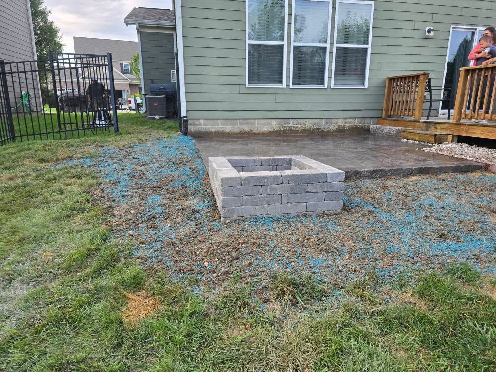 A backyard with a fire pit made of gray blocks on a concrete patio and patchy grass.