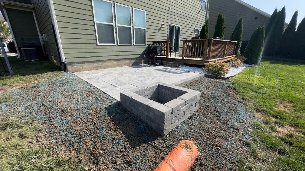 Backyard with stone fire pit, patio, wooden deck, and a house. Green grass and blue sky.