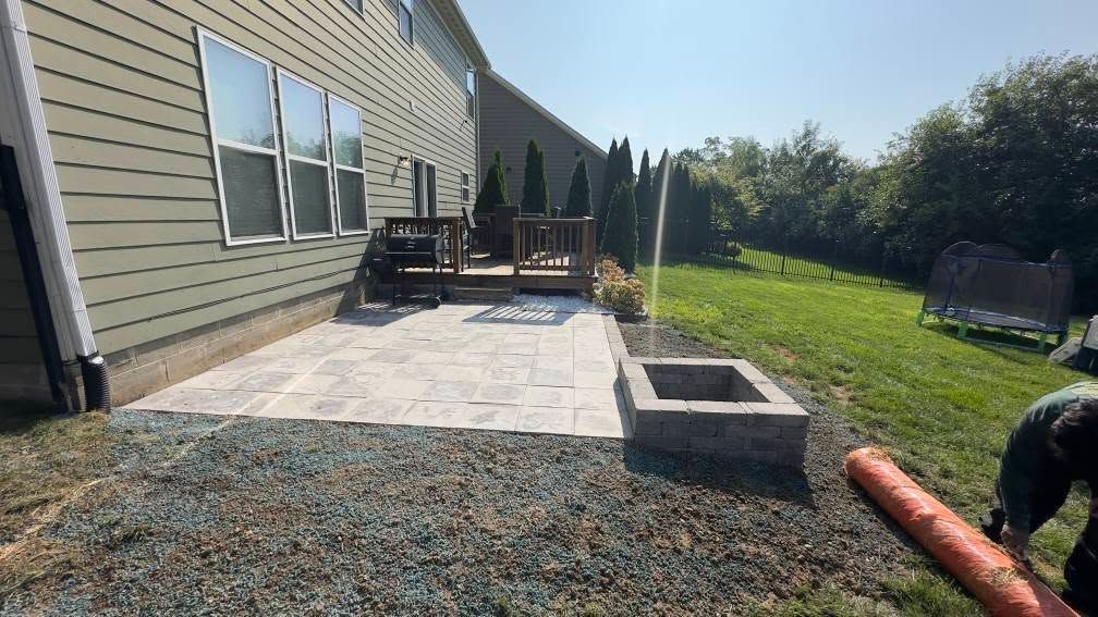 Backyard patio with fire pit, deck, and house. A person works on the ground.