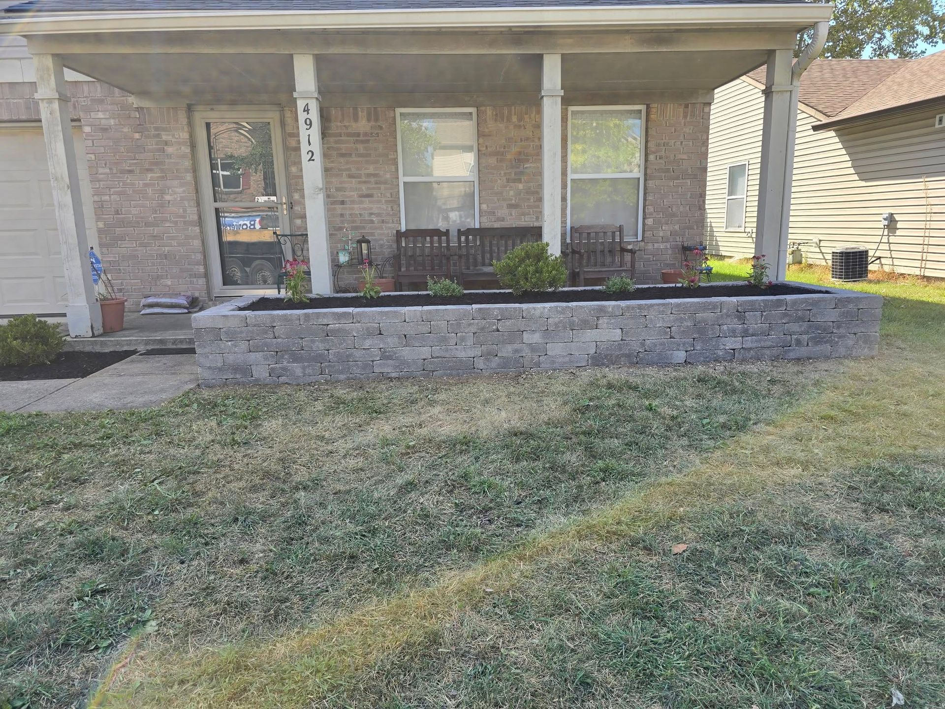 Low-angle view of a house with a raised stone planter. Front yard has dry grass. Porch with two windows.