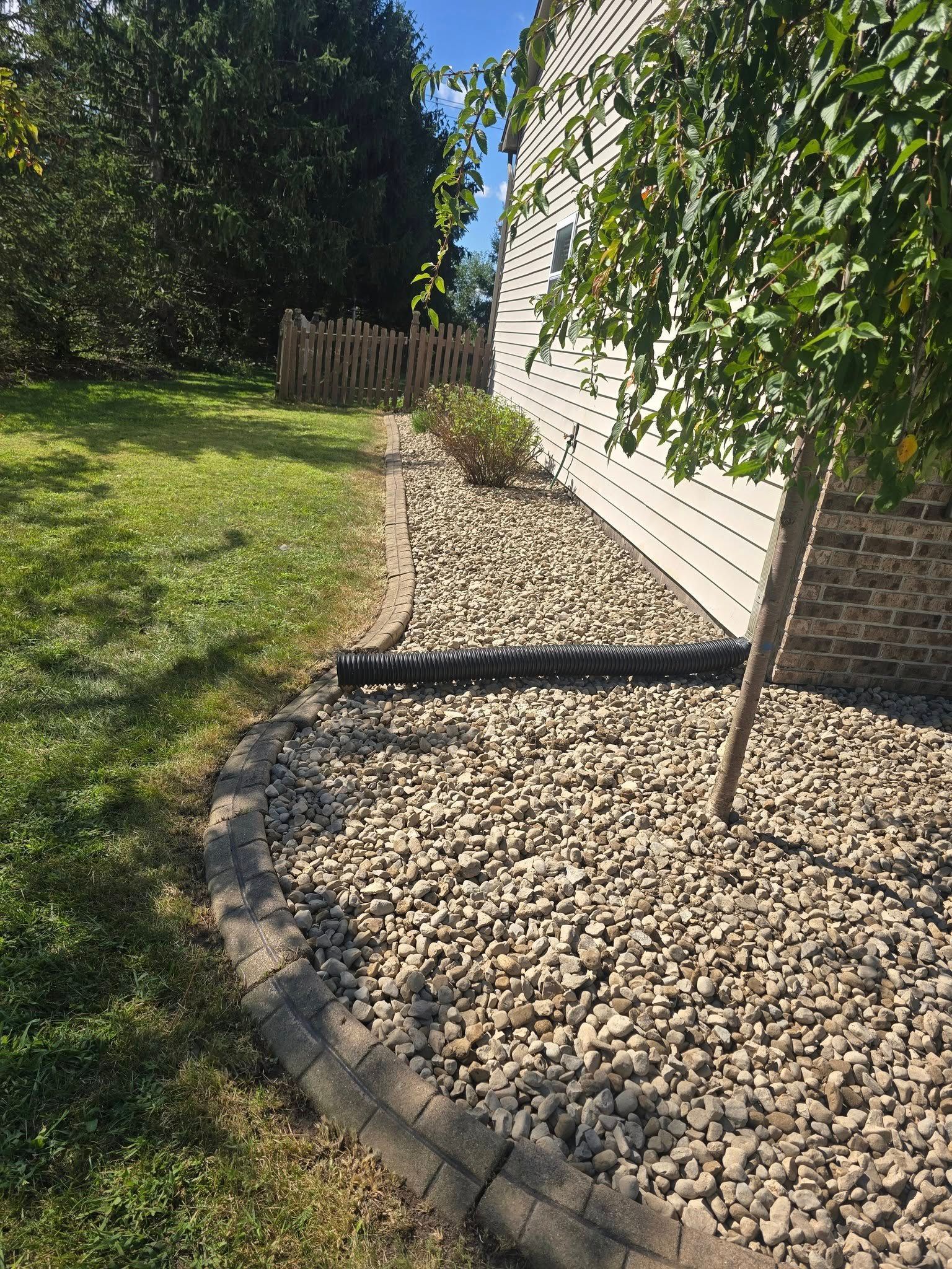 Rocks and landscaping border along the side of a house with a grassy yard and small fence.