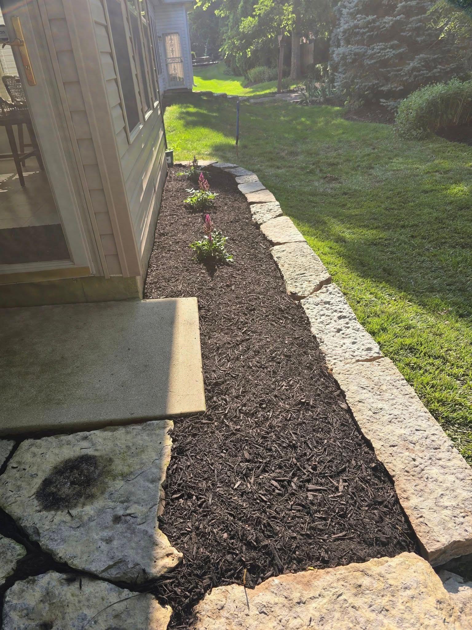 Stone patio next to a narrow mulch bed with a stone border. The sun is shining.