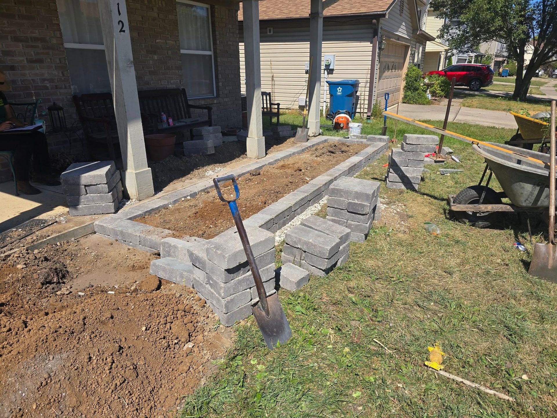 Construction of a brick planter box near a porch. A shovel rests on the soil.