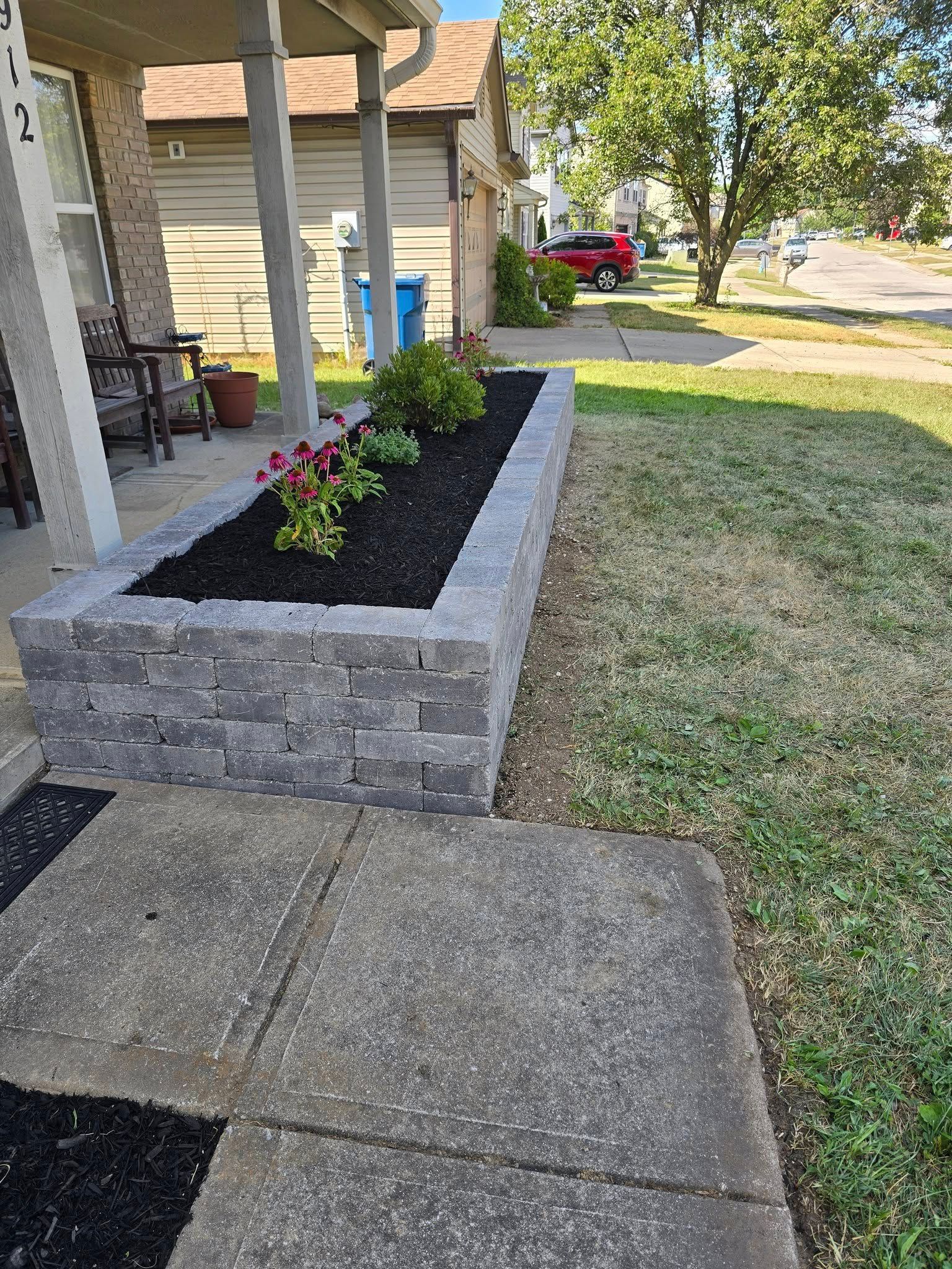Raised flower bed made of gray blocks with flowers and mulch, next to a concrete walkway.