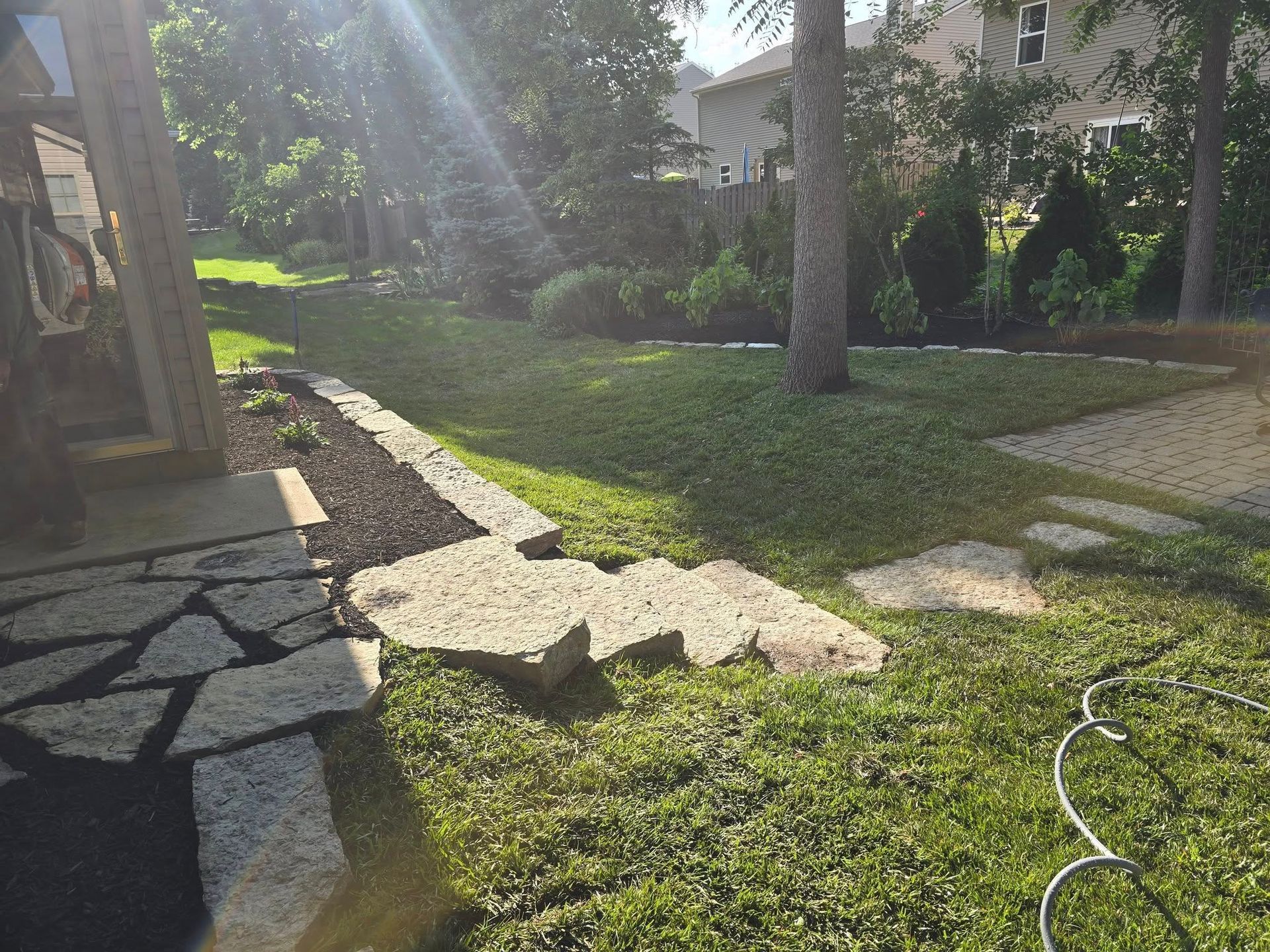 A sunlit backyard with stone path, steps, and landscaping. Trees and grass are visible.