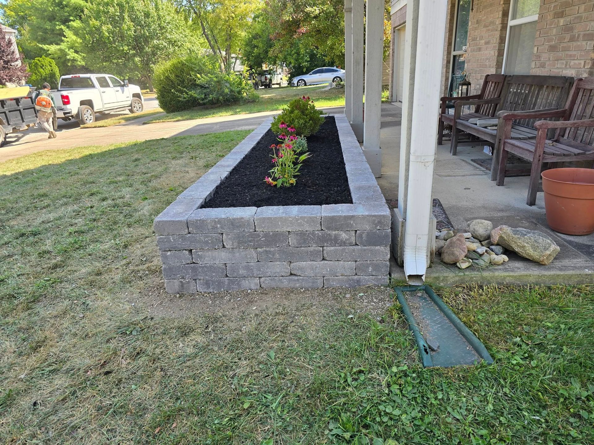 Raised garden bed with gray retaining wall blocks, containing dark mulch and flowers, in front of a porch.