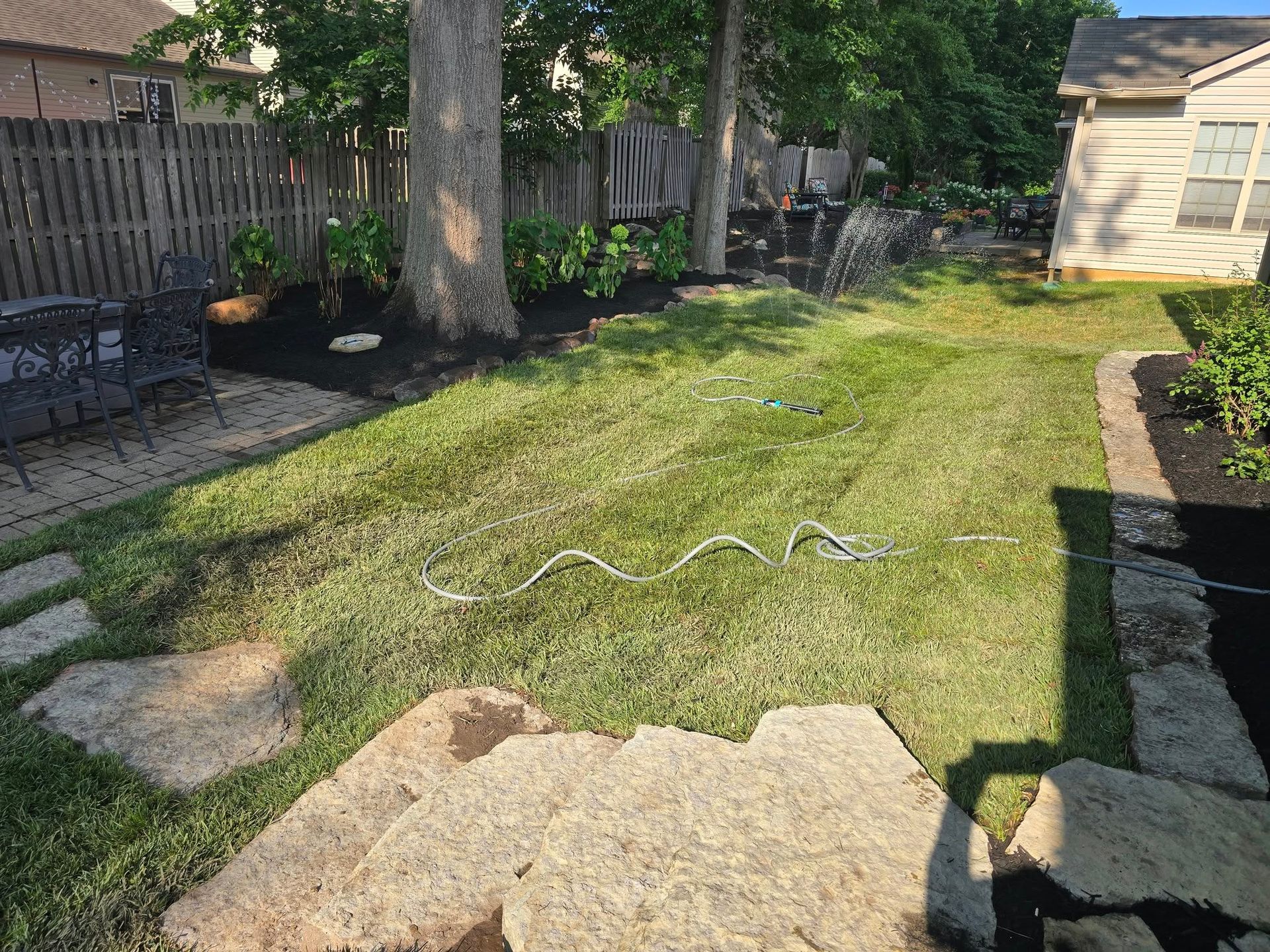 Backyard with stepping stones, lawn, mulch, trees, waterfall, and fence.