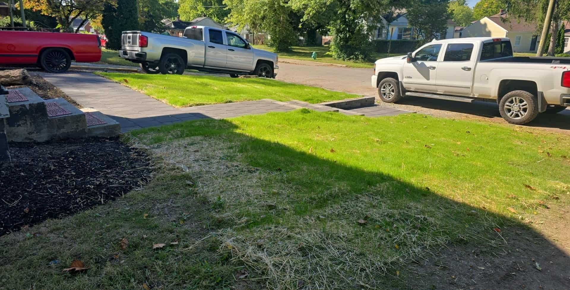 Three pickup trucks parked on grass and a street in front of houses; sunny day.