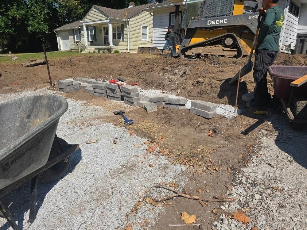 Construction site with a worker, wheelbarrow, tractor, and retaining wall blocks.