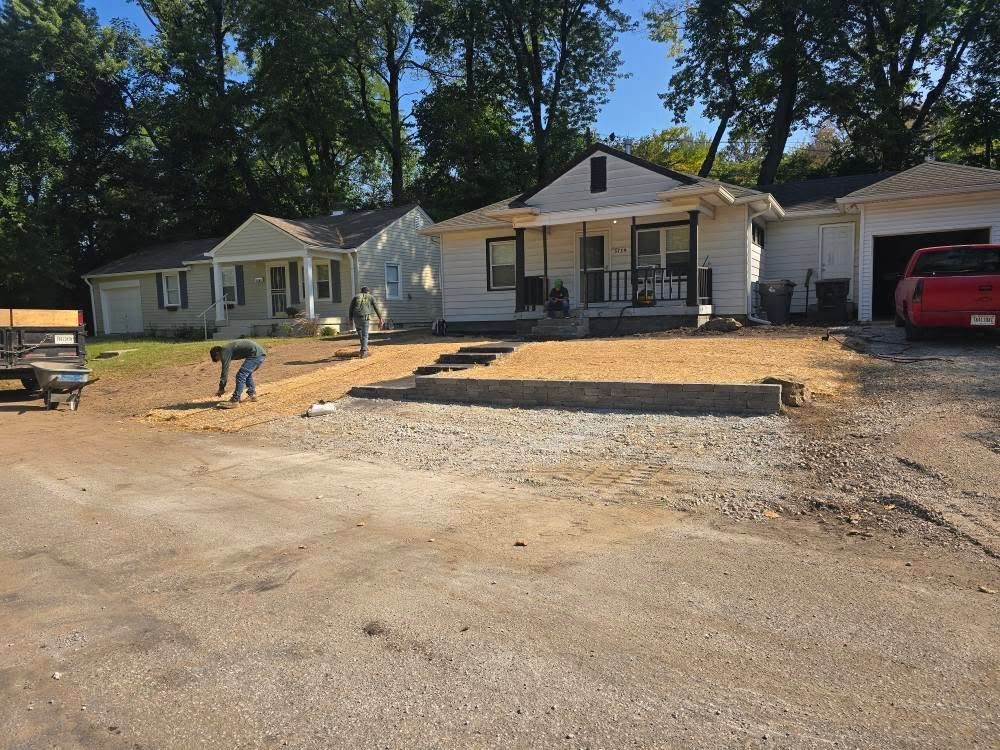 Houses and gravel area; two people work on a gravel border in front of a house, with a truck parked nearby.