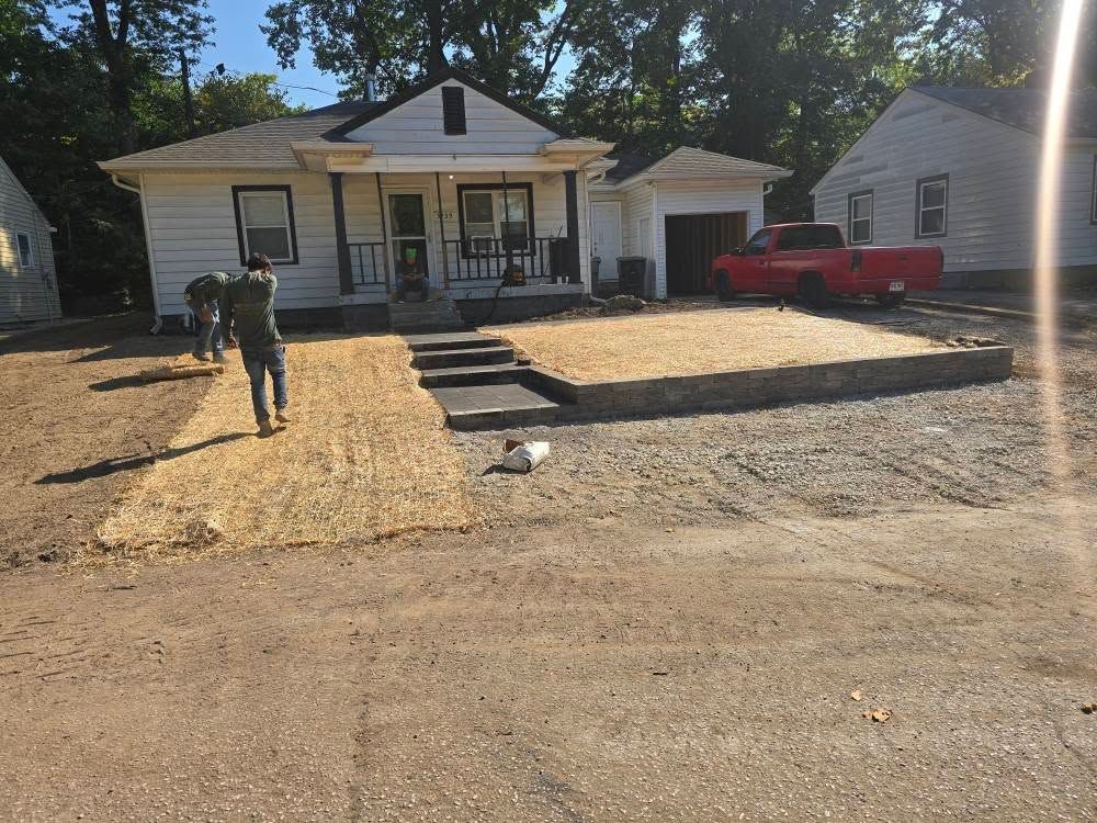 House with dirt yard and workers, gravel driveway. A red truck is parked in the garage.
