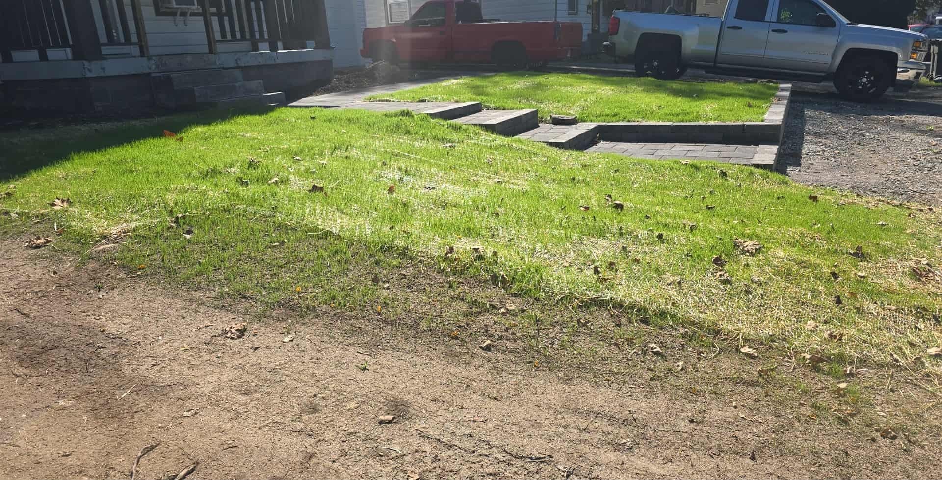 Green lawn and concrete structure next to a dirt driveway, with a house and vehicles in the background.