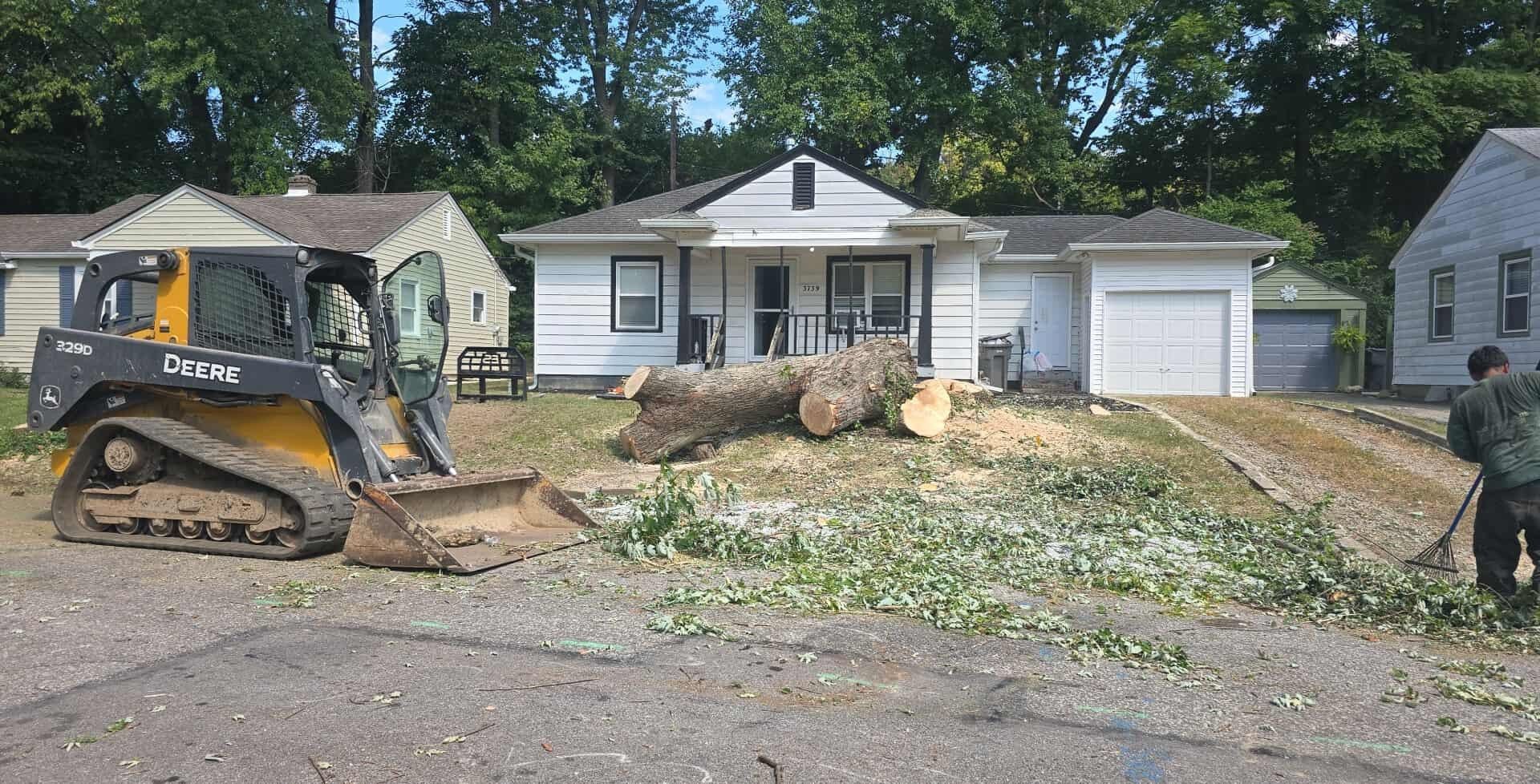 Tree removal in progress: A skid steer and workers near a house; debris and a large log are on the lawn.