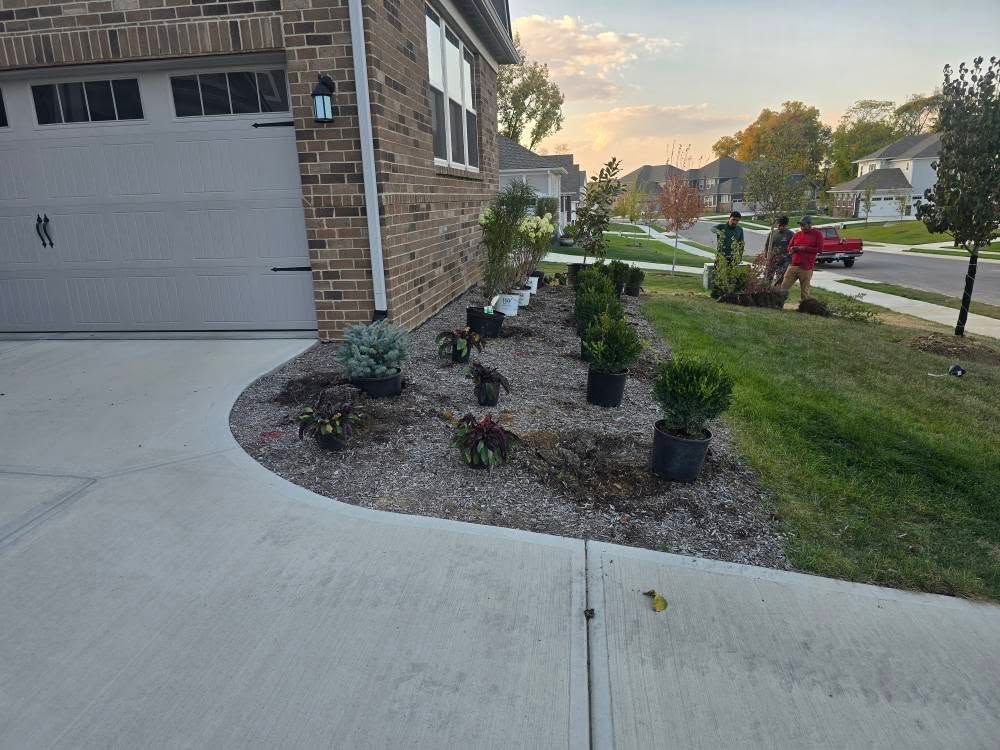 Landscaping in progress: Plants in pots near a house, gravel bed, driveway, people working, cloudy sky.