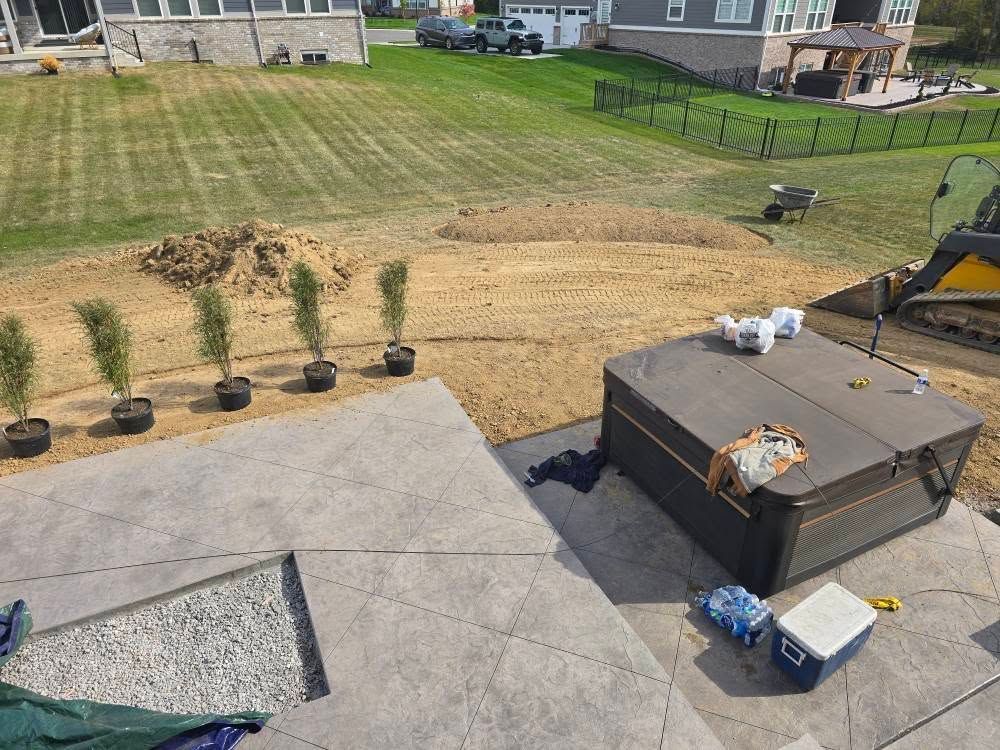 Backyard with hot tub on a concrete patio. Trees in pots, grading underway, and vehicles in the distance.