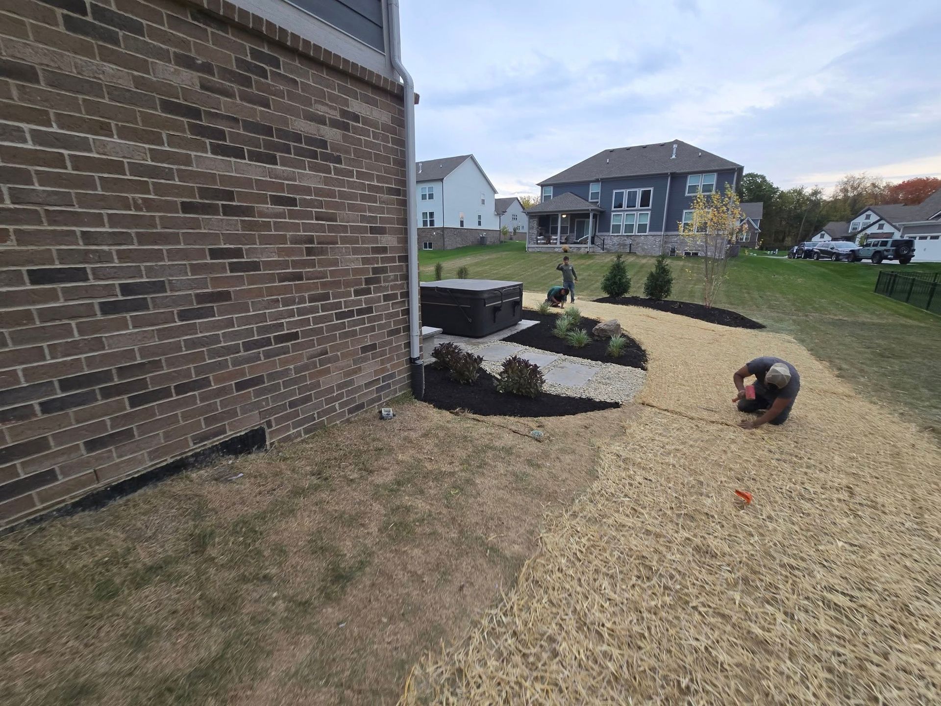 Backyard landscaping: brick house, mulch, bushes, HVAC unit, person working. Sunny day.