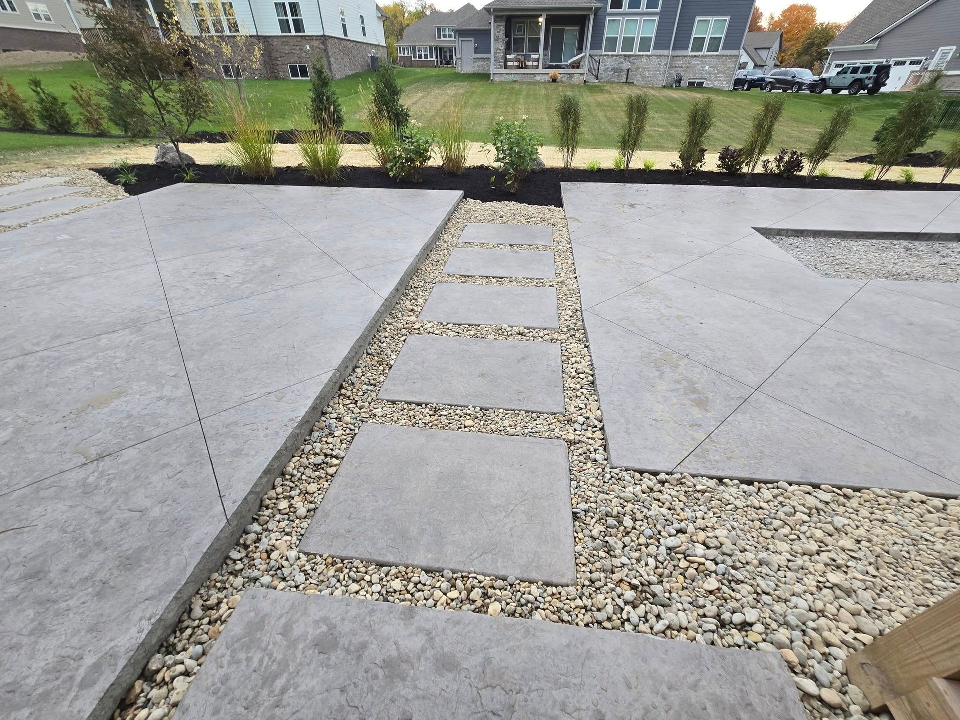 Gray stone patio with stepping stones through gravel, and landscaped border in a backyard.