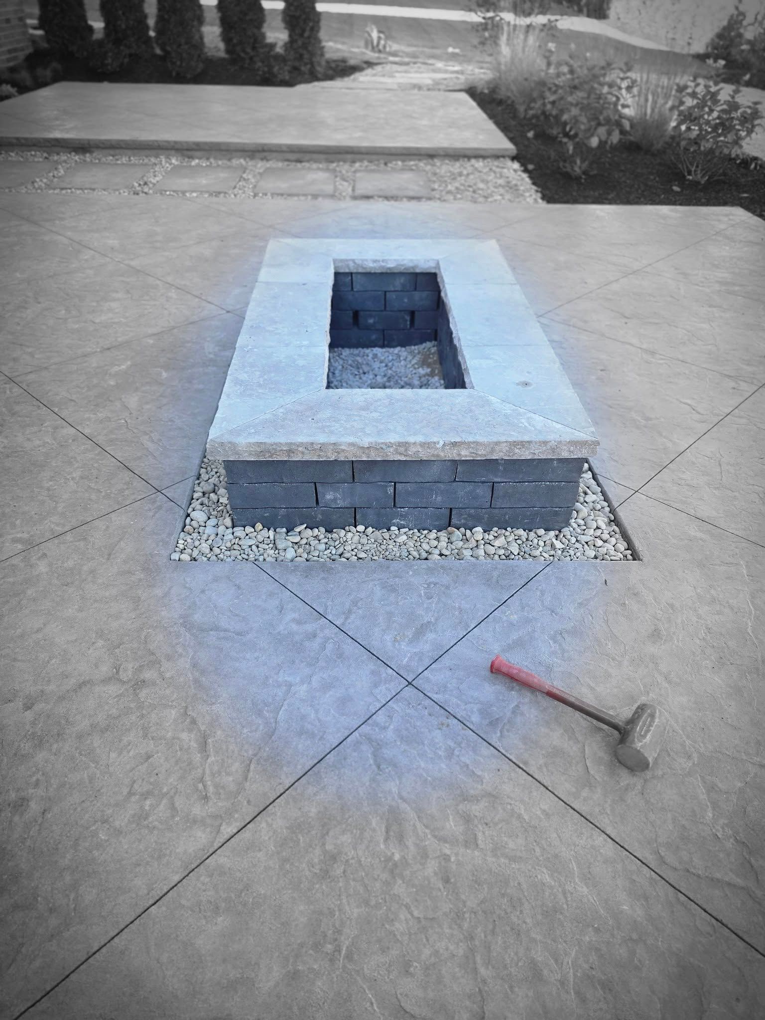 Rectangular stone fire pit on a gravel base within a gray concrete patio, a hammer nearby.