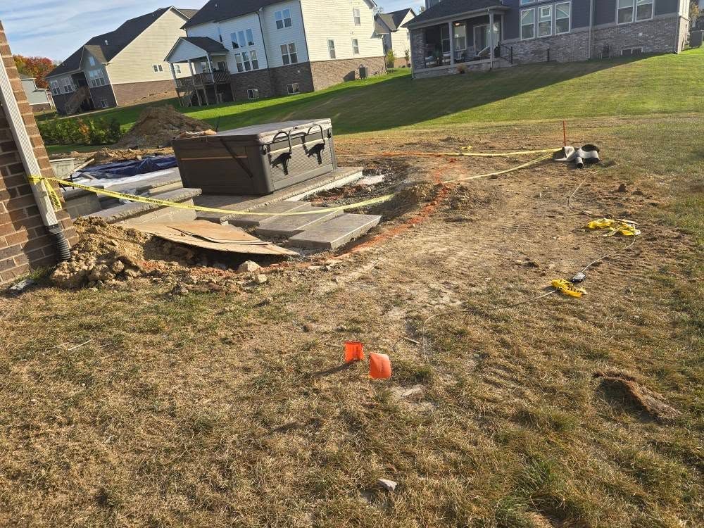 Backyard construction site: earth moved, stone slabs, brown box, caution tape, yellow and orange markers, grassy hill, houses in background.