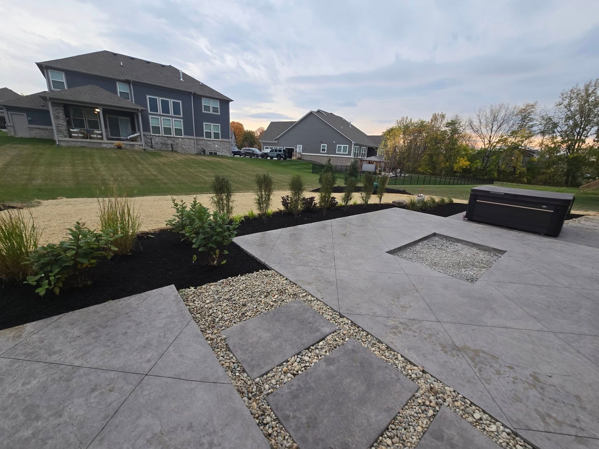 Backyard patio with landscaping, concrete pavers, and a hot tub. Houses in the background under a cloudy sky.