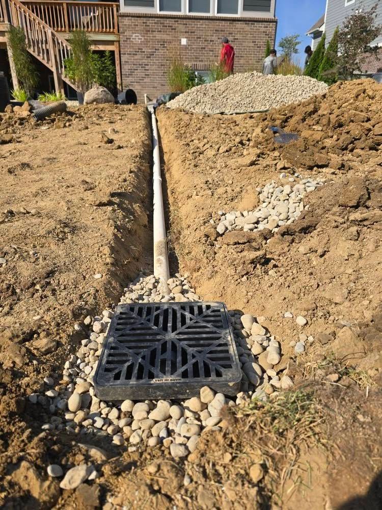 Drainage system under construction in a yard. Black grate in dirt, connected to a pipe leading uphill. Gravel and a person in the background.