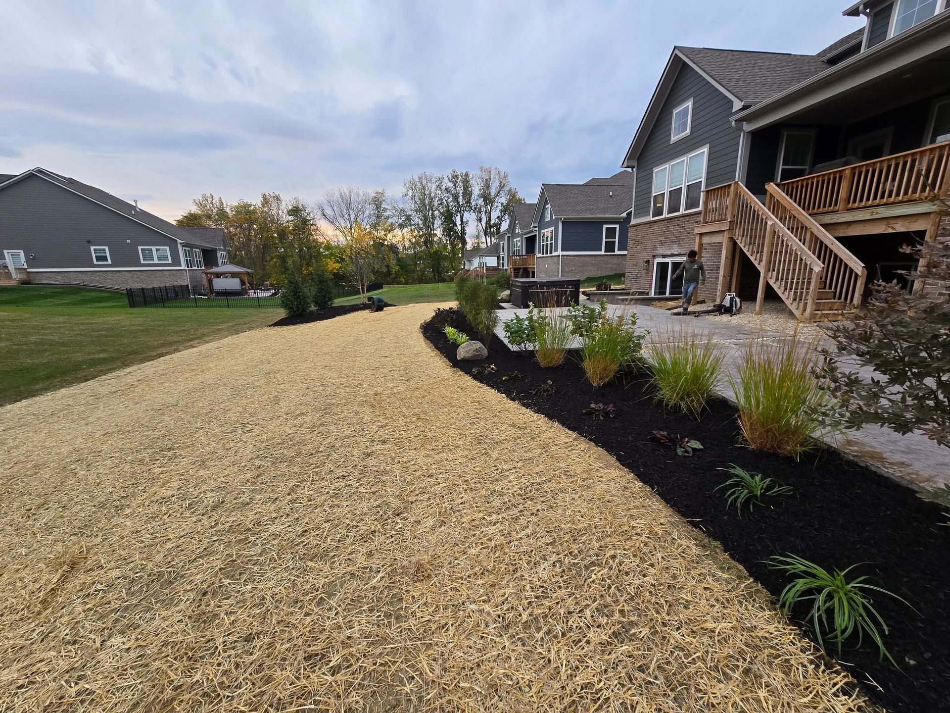 Backyard landscaping with mulch pathway, flowerbeds, and houses in the background. Cloudy sky.