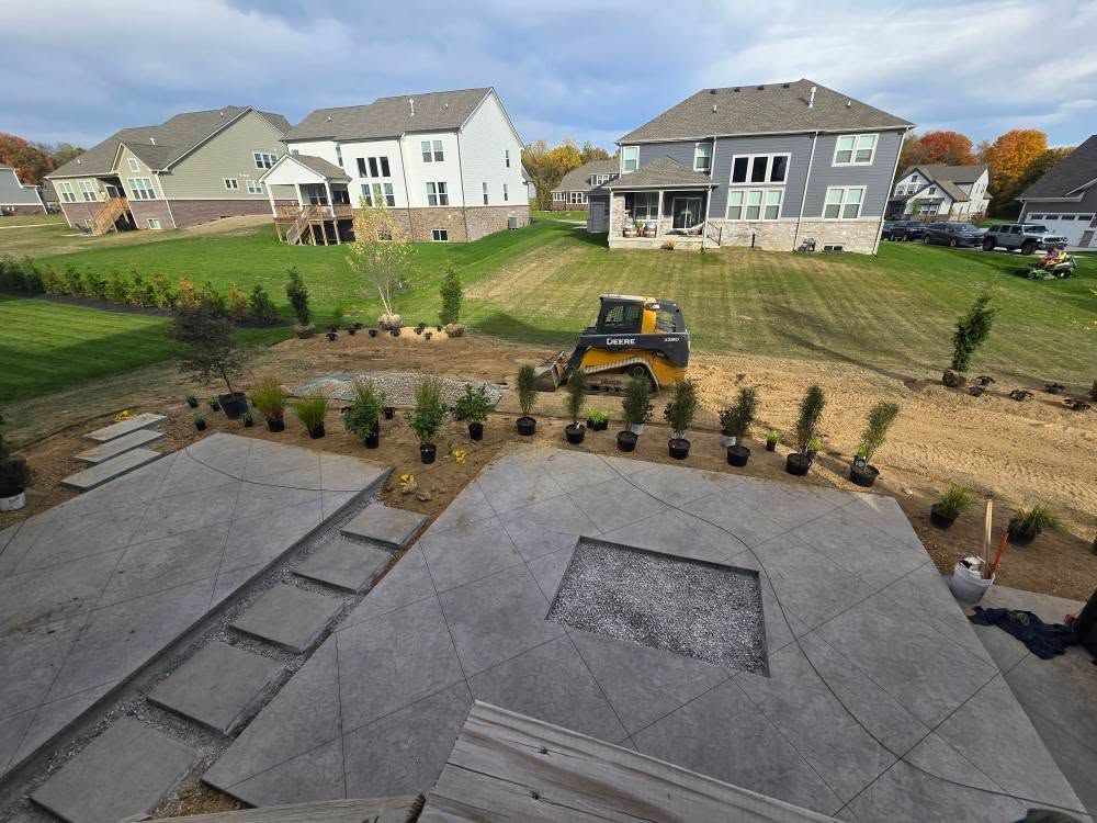 Construction site with a concrete patio, steps, and plantings, a skid steer, and houses in the background.