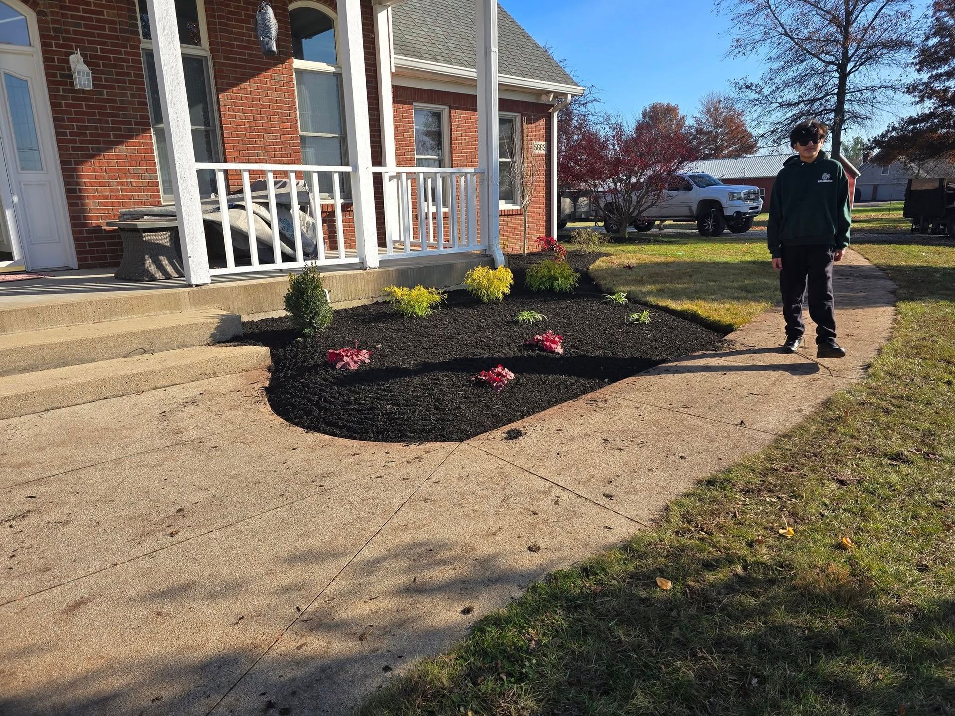 Person standing next to a landscaped flower bed in front of a brick house with a white porch.