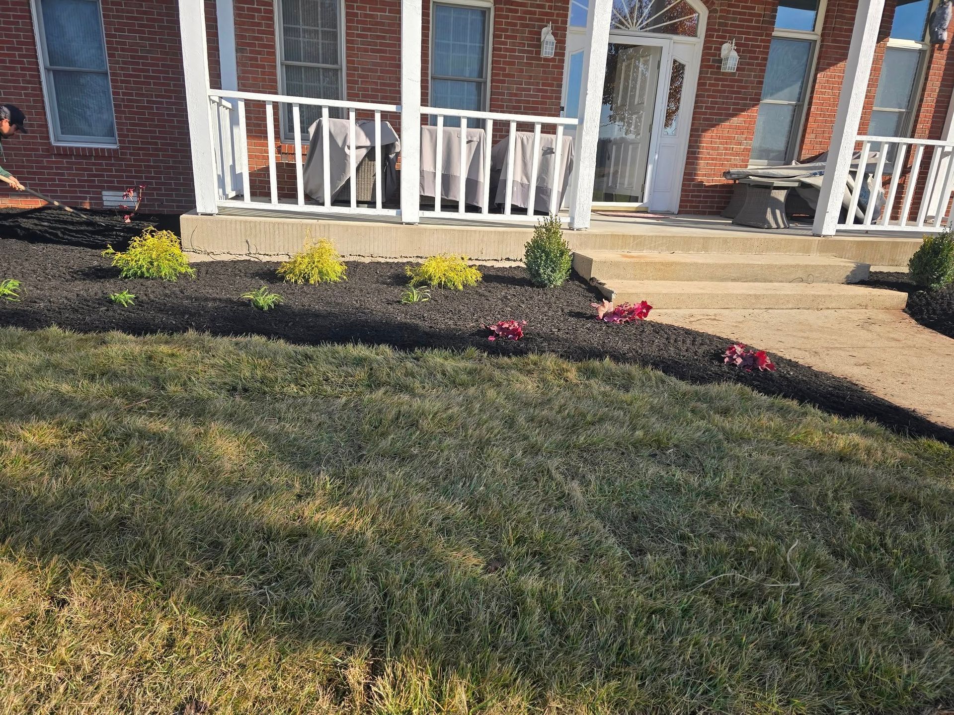 Front yard with brick house, porch, steps, dark mulch bed, green grass, and small plants.