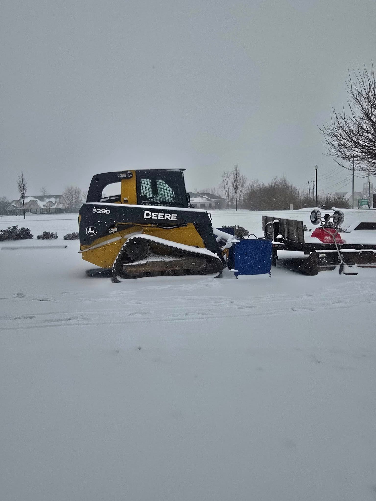 Yellow and black skid steer in the snow, with a trailer in the background.