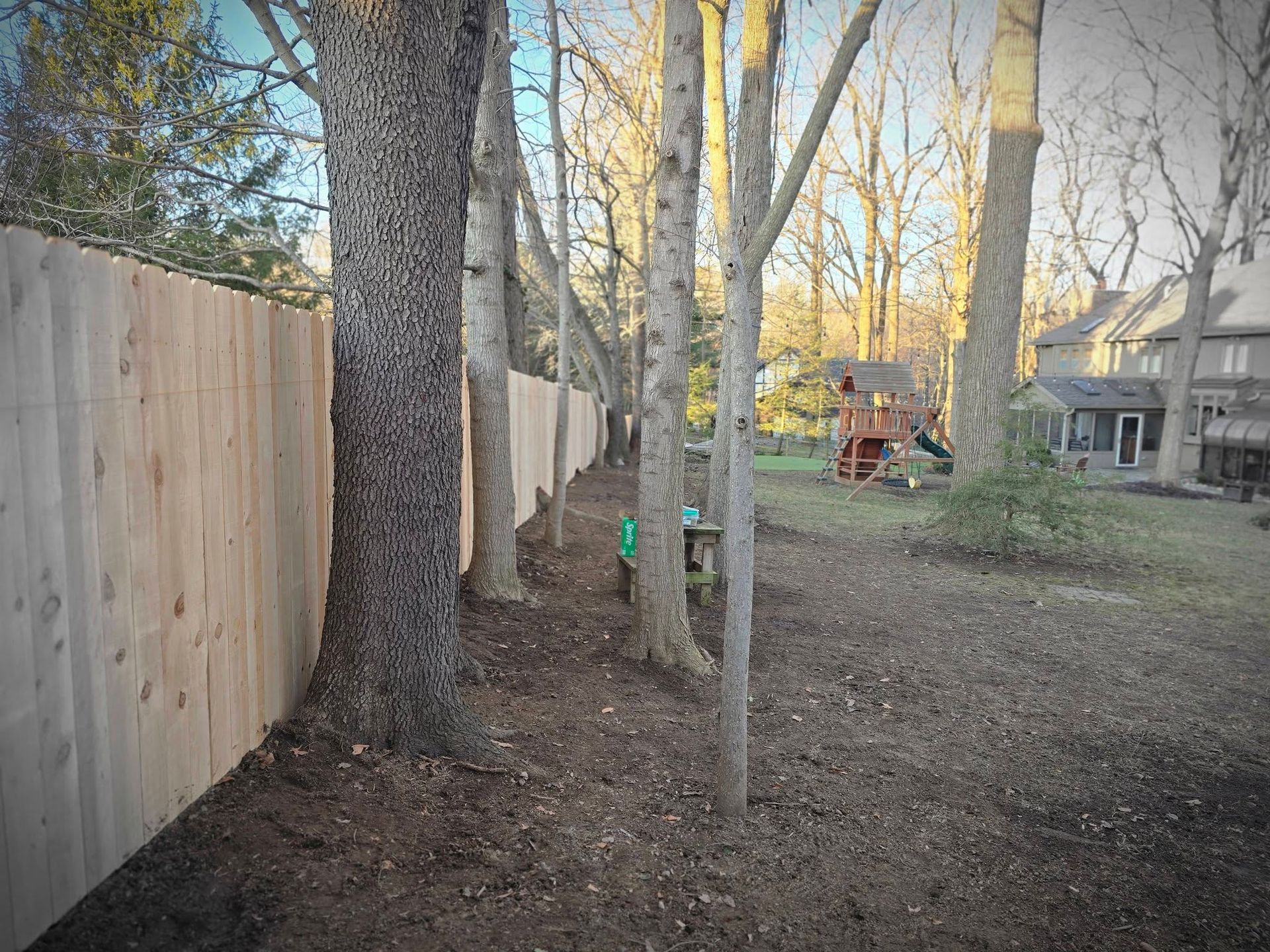 Wooden fence lines backyard, trees in between, with a play set and house visible in the distance.