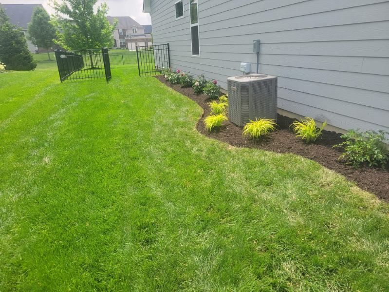 Green lawn with a flower bed and house exterior. Mulch bed with flowering plants, air conditioner, and fence.