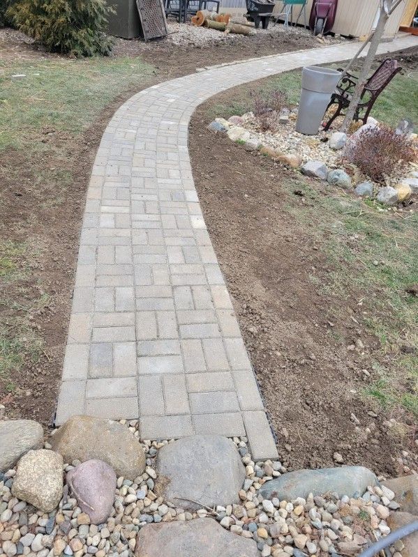 Brick pathway curves through a yard with rocks and gravel along edges; grass on either side.