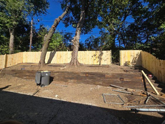A newly built wooden fence surrounds a dirt yard with two trees. Construction materials are scattered about.