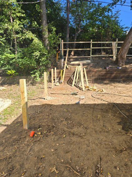 Construction site: wooden fence posts partially installed in dirt, tools and a partially built fence in background.