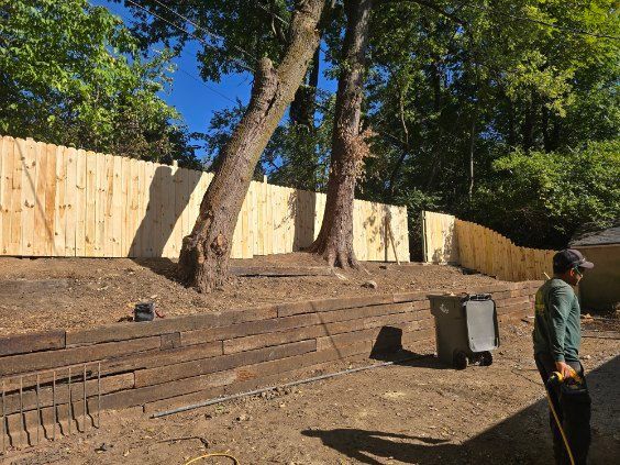 Newly built wooden fence runs along a slightly elevated yard with trees and a worker standing nearby.