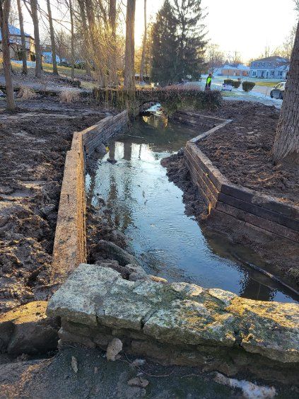 A channelized stream with wooden retaining walls, a bridge in the distance, and a person in a vest.