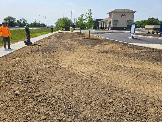 Two workers near a newly prepared, dirt-filled area beside a building with a drive-thru, paved lot.